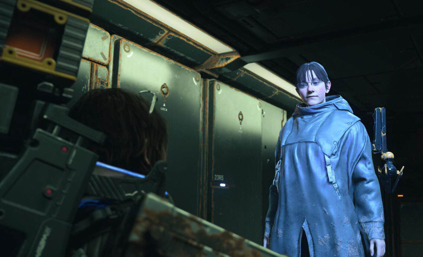 A woman with glasses and a blue raincoat looks at a man inside a corridor of rusty metal containers.